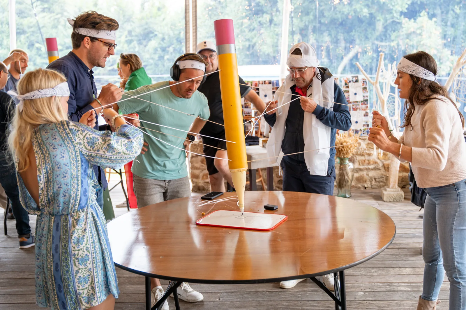 Équipe en bandeaux blancs manœuvrant un crayon géant autour d'une table, animation en terrasse