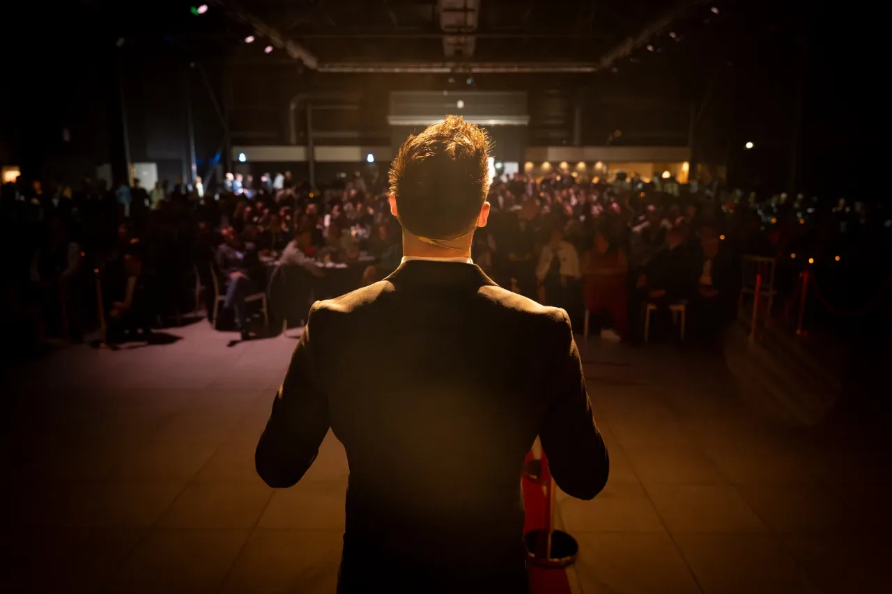 Conférencier vu de dos face à une salle comble plongée dans la pénombre