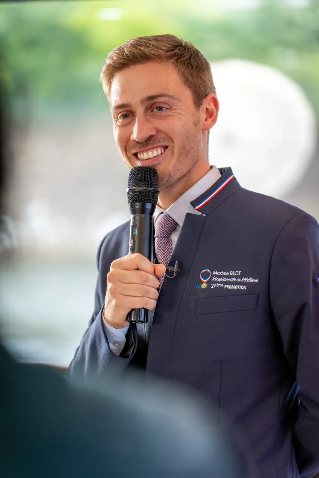 Intervenant souriant, micro en main, devant une grande baie lumineuse